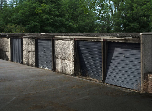 battery-dismantle - Hanson Battery Garages Row of old metal garage doors in a concrete wall, with green trees behind them by a paved lot.